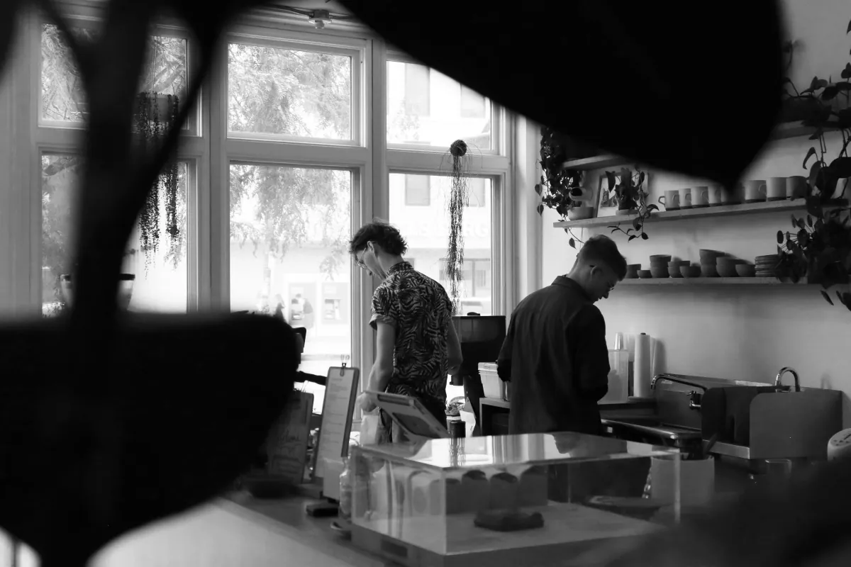 Two baristas at work behind the Persimmon counter, seen in black and white through the pastry case, with mugs and bowls on wall shelves and the street beyond the window