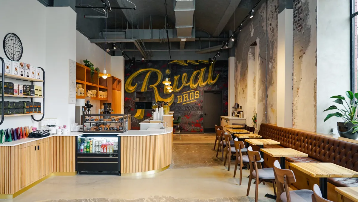The Brewerytown café of Rival Bros, with a curved slatted-oak counter, a pastry case, a yellow Rival Bros mural on a dark wall and a leather banquette along the right