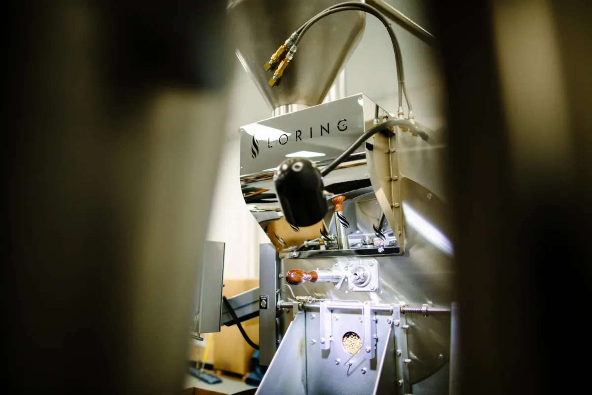 The polished steel drum and hopper of Vibrant's Loring roaster, seen between two blurred figures in the foreground