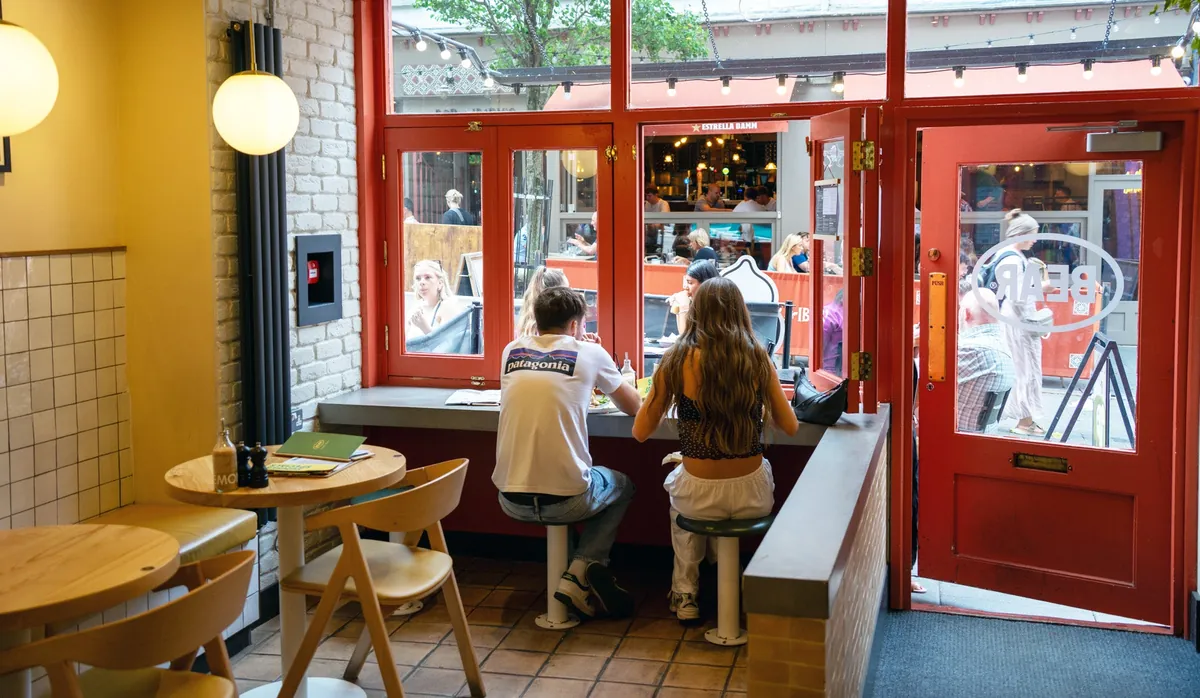 Customers at a window counter inside BEAR Hockley, framed by red doors and windows, with small wooden tables and globe lamps