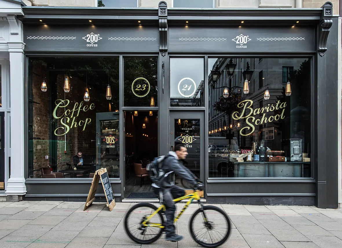 The dark grey shopfront of 200 Degrees below the Grand Hotel in Birmingham, with Coffee Shop and Barista School painted on the windows and a cyclist passing