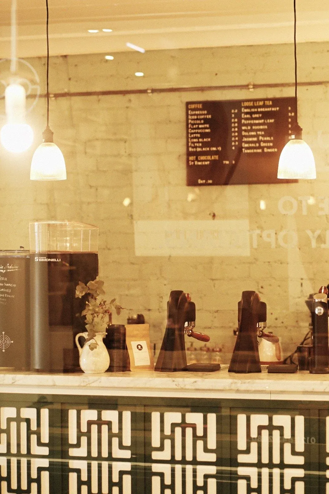 The counter at Faculty Coffee seen through the window, with two lever-handled grinders, a batch brewer and a coffee and tea menu board on a white brick wall
