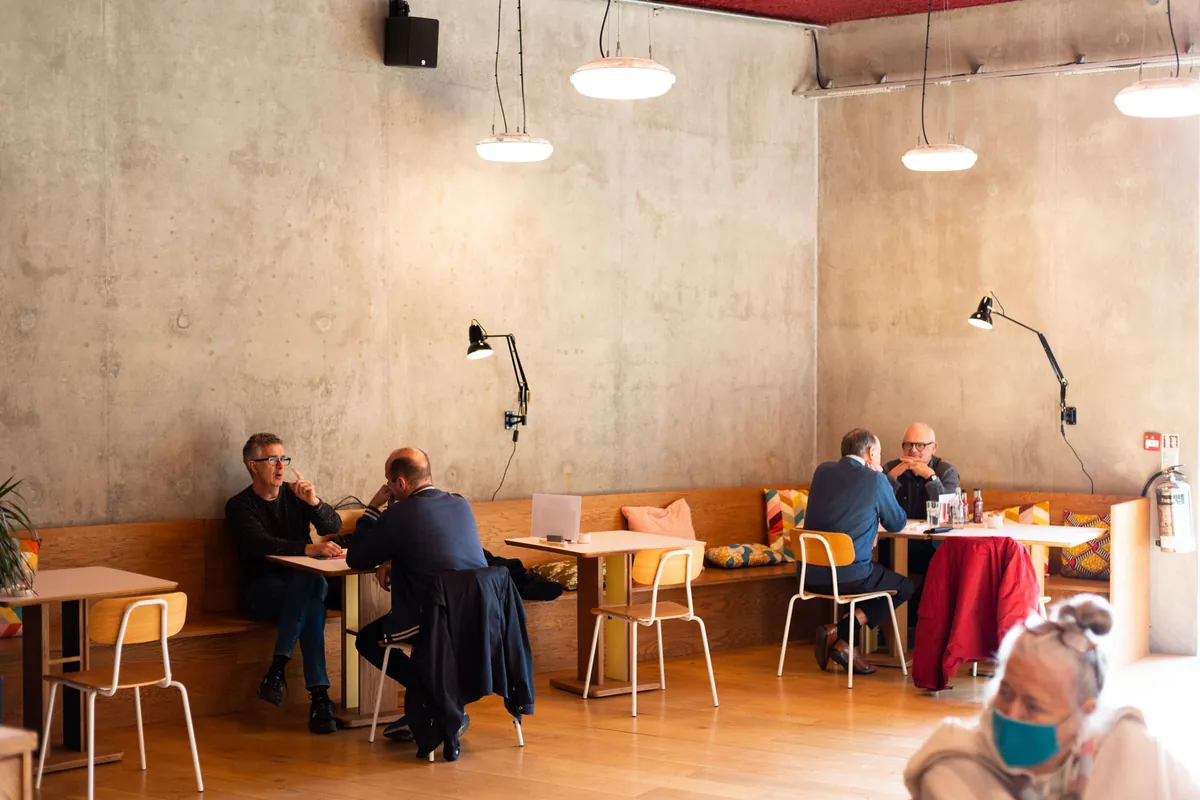 Customers at small wooden tables in a Blend coffee shop, with bare concrete walls, pendant lamps and a long bench along the wall
