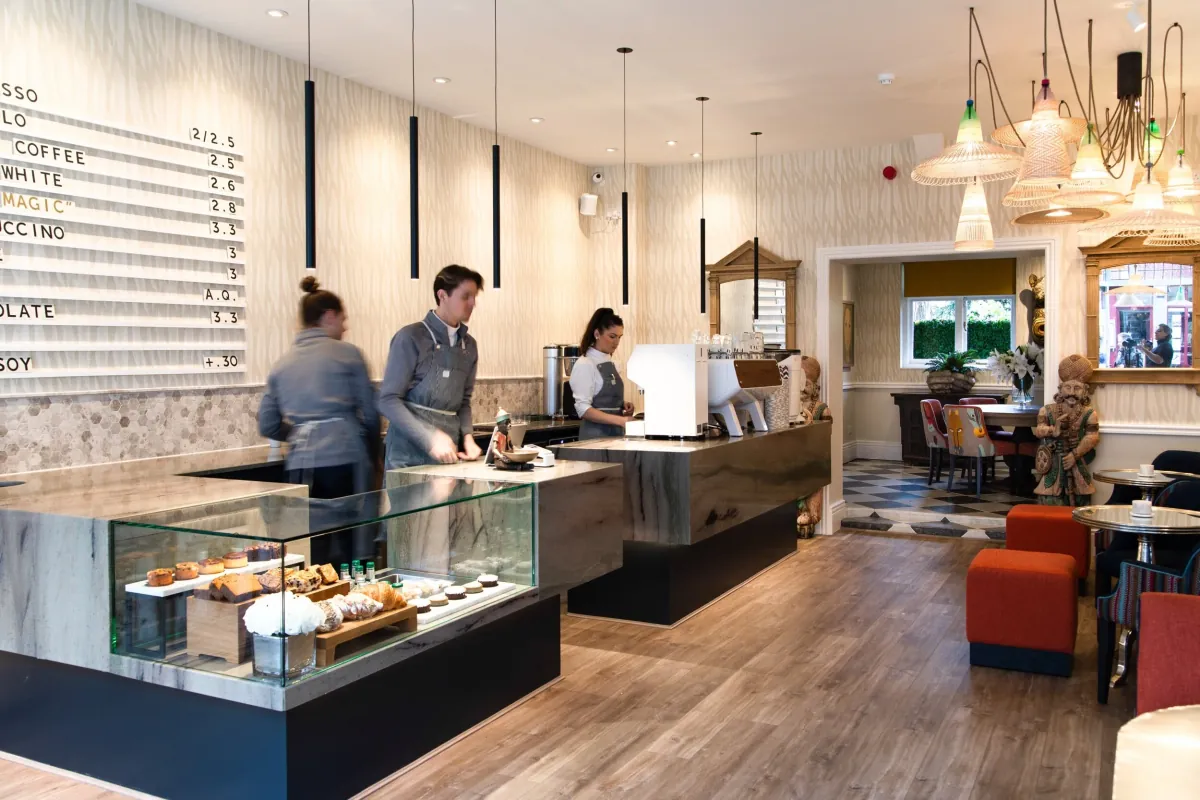 Baristas behind the long counter at Bould Brothers in Cambridge, with a pastry case, a menu board and orange armchairs at the back
