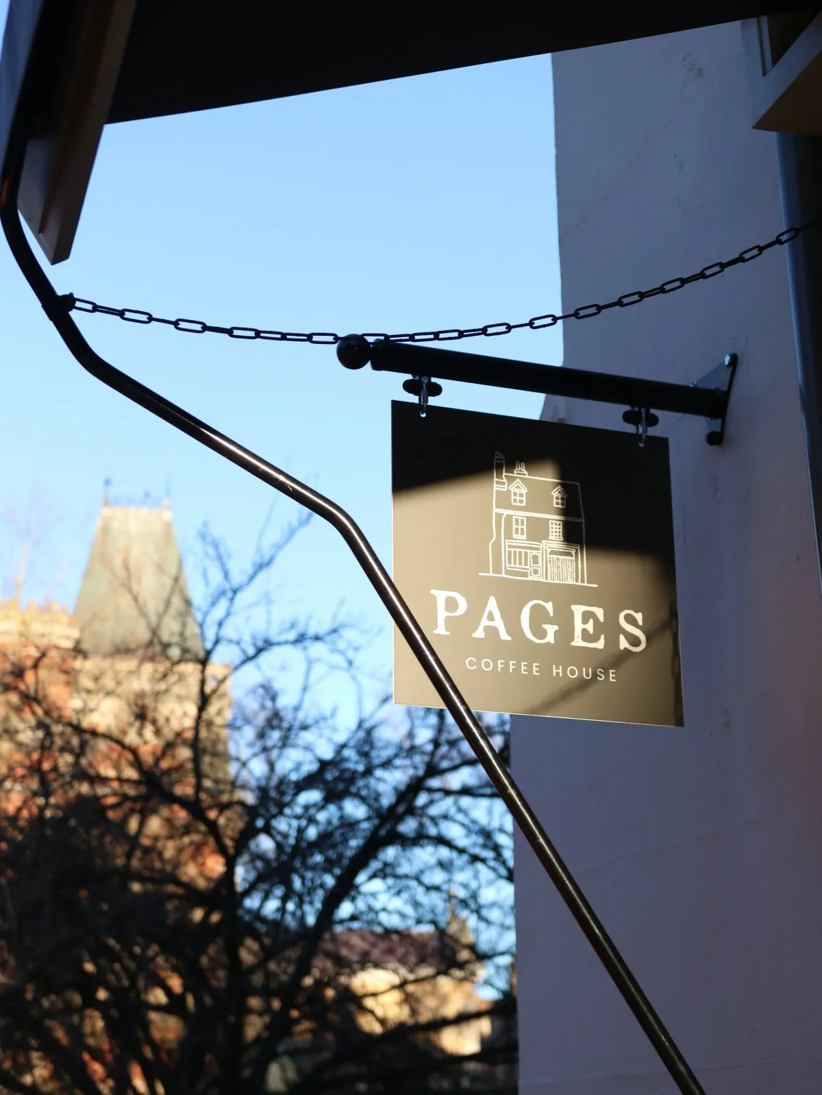 The hanging sign of Pages Coffee House against a blue sky in central Cambridge, with a stone tower behind