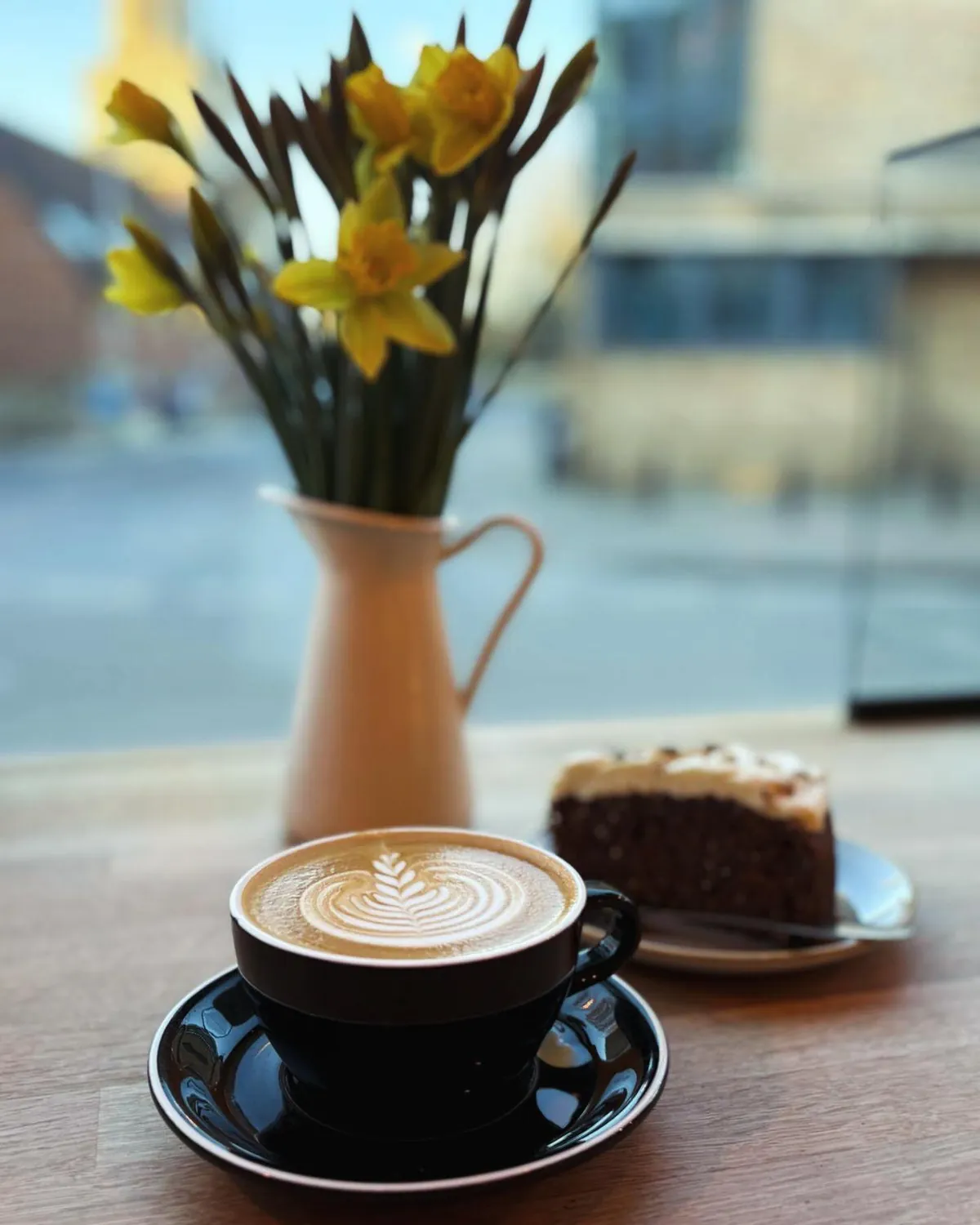 A flat white with latte art and a slice of cake on a window table at The Locker Cafe in Cambridge, beside a jug of daffodils