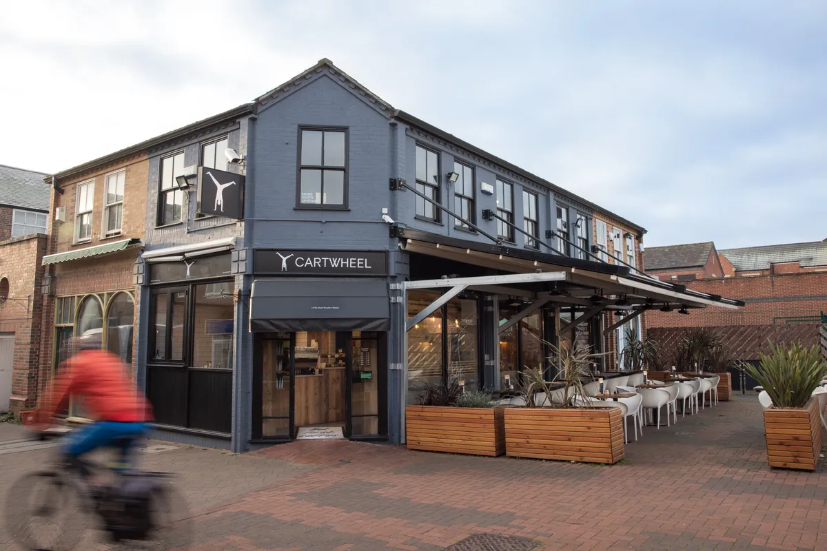 The slate-blue corner building of Cartwheel’s Beeston café, with the CARTWHEEL sign over the door, a covered terrace with planters and a cyclist passing