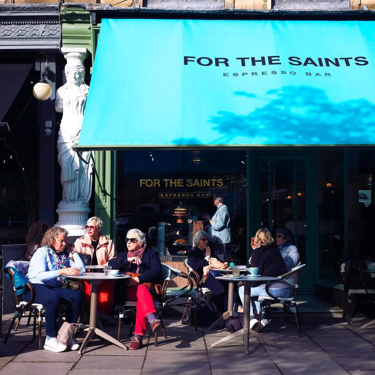 Customers at café tables under the turquoise For The Saints Espresso Bar awning in Montpellier, Cheltenham, beside a white caryatid statue