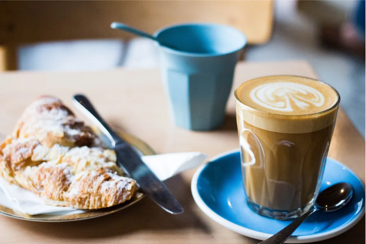 A flat white in a glass on a blue saucer beside an almond croissant on a café table at Suffolk Coffee in Cheltenham