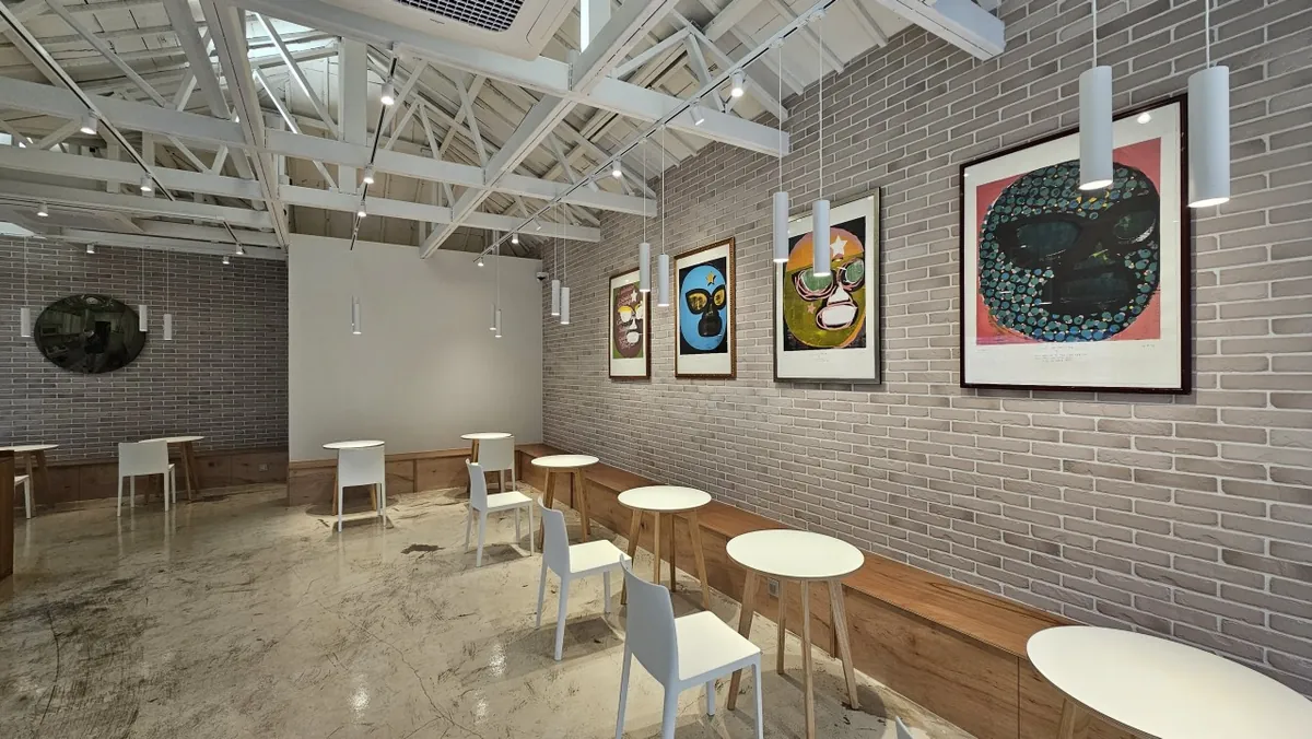 The seating area of Coffee Libre Yeonnam, with white chairs and round tables under a white steel roof truss and framed luchador-mask prints on a grey brick wall