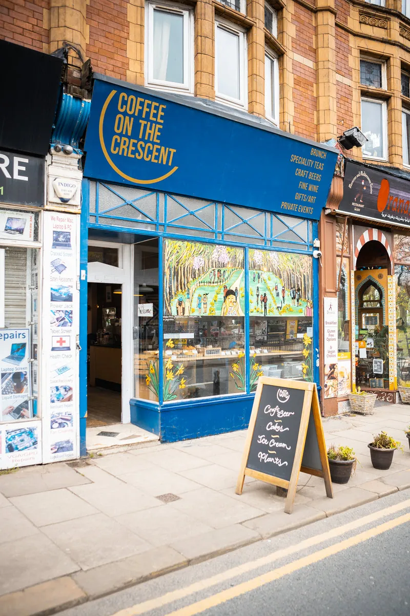 The blue and glass shopfront of Coffee On The Crescent in Hyde Park, Leeds, with a chalkboard sign and plants on the pavement