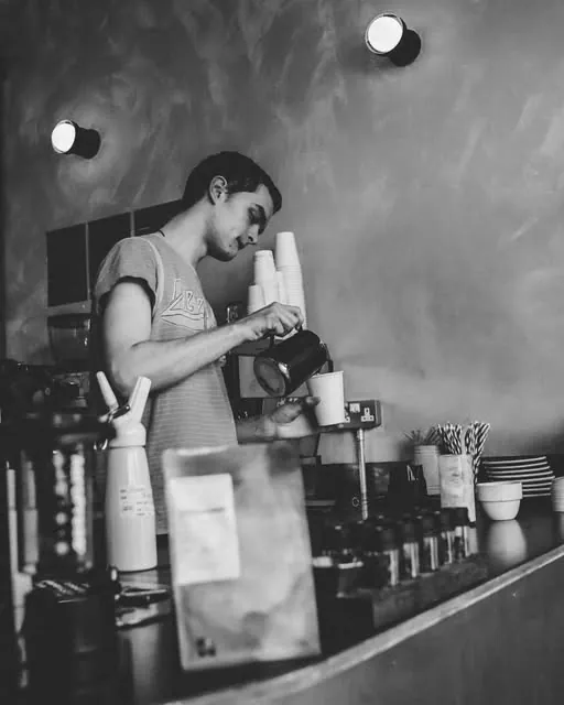 A barista steaming milk at the espresso machine behind the bar at hang ten. brew bar, in black and white