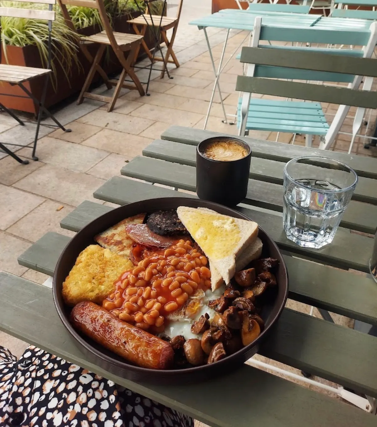 A cooked breakfast with sausage, beans, mushrooms and toast beside a coffee on a green slatted table on the terrace at Nine Hostages