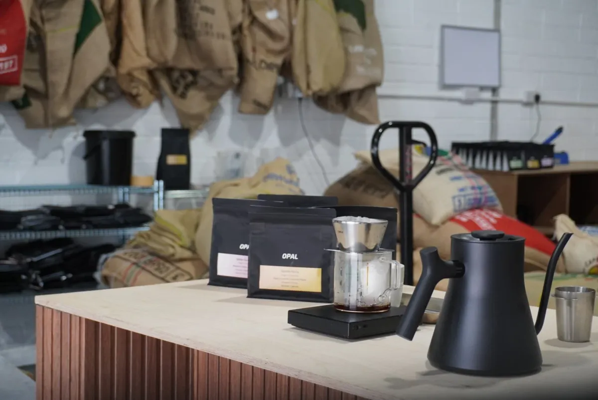 A pour-over brewer, a black gooseneck kettle and Opal coffee bags on a wooden counter, with green coffee sacks hanging on the wall behind