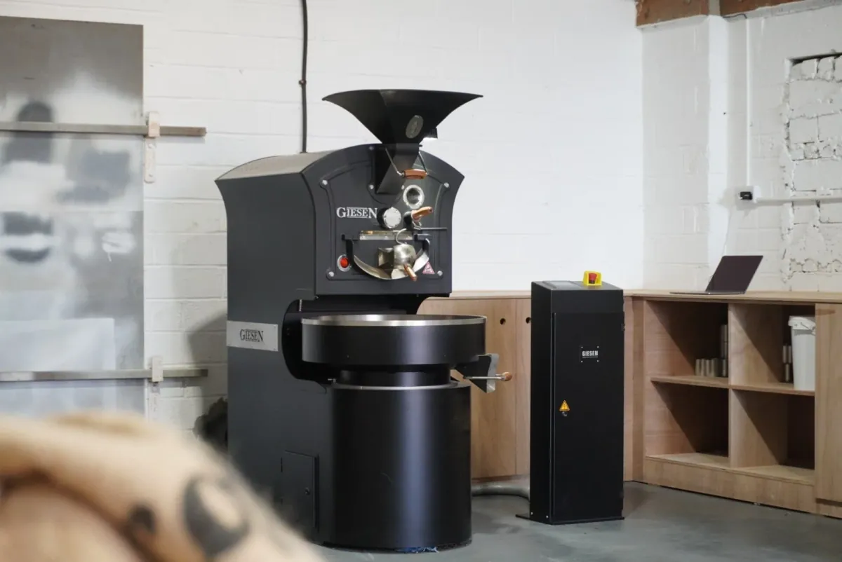 A black coffee roaster beside wooden shelving in the Opal roastery, with a sack of green coffee in the foreground