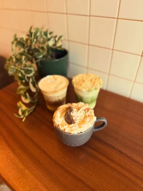 A toffee apple latte, a Terry's hot chocolate and a cheesecake matcha on a wooden table against white tiles at The Coffee Tree