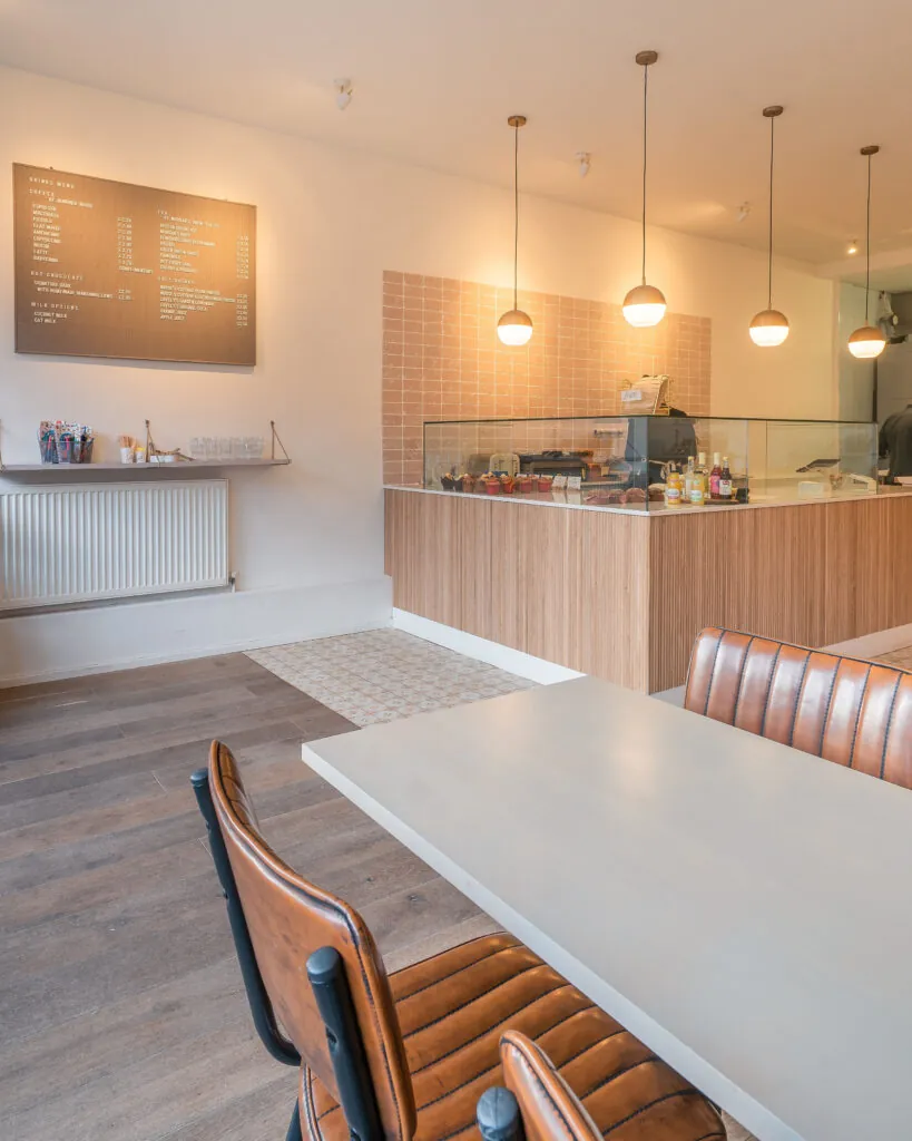 Inside The Early Bird Bakery in Kings Heath, with leather chairs at a pale table, globe pendant lights and a wooden counter with a cake cabinet under a pink tiled wall