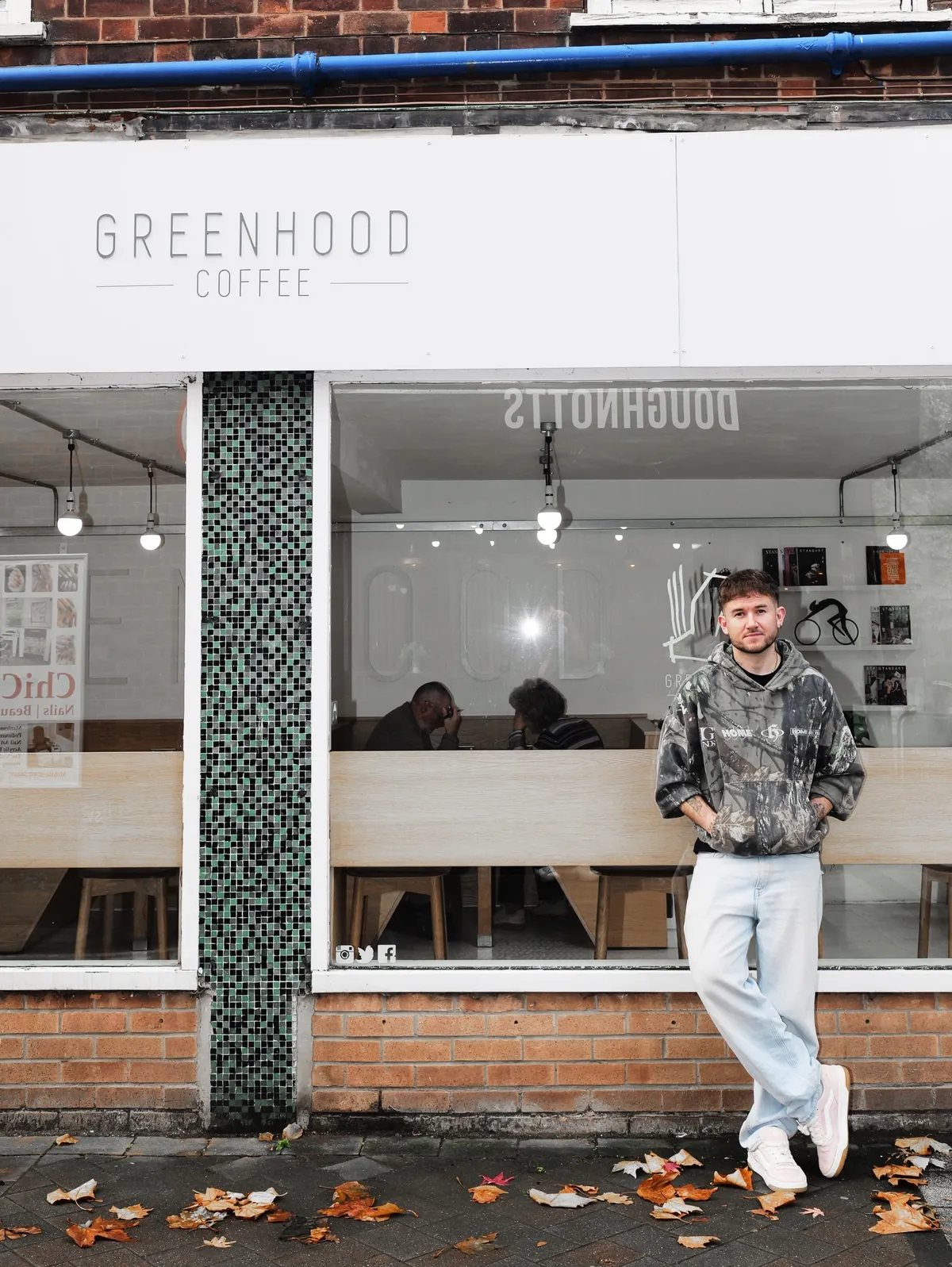 Greenhood's owner standing outside the white Greenhood Coffee shopfront in Beeston, with a green tiled pillar and autumn leaves on the pavement