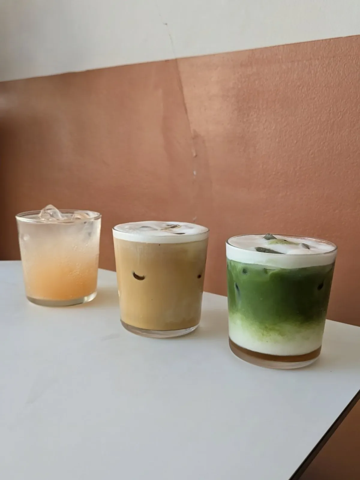 Three iced drinks, among them an iced latte and an iced matcha, on a white table against the terracotta wall at Esters
