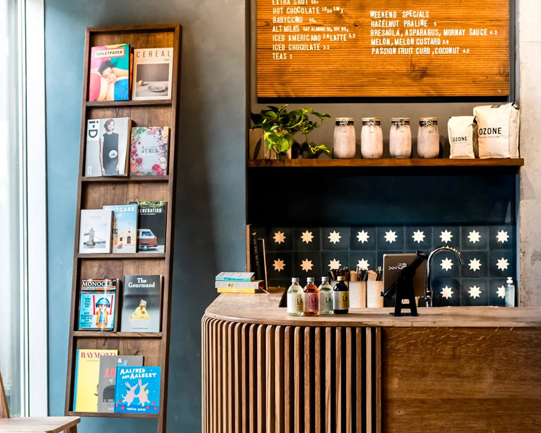 The counter at Pophams London Fields, with a curved wooden front, star-patterned tiles, a wooden menu board and a rack of magazines