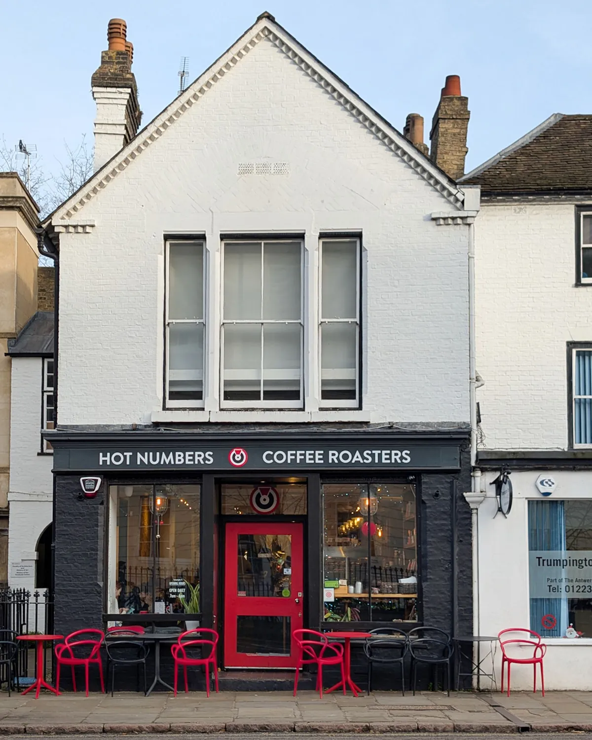 The white gabled front of the Hot Numbers city-centre coffee shop in Cambridge, with a red door and red chairs on the pavement