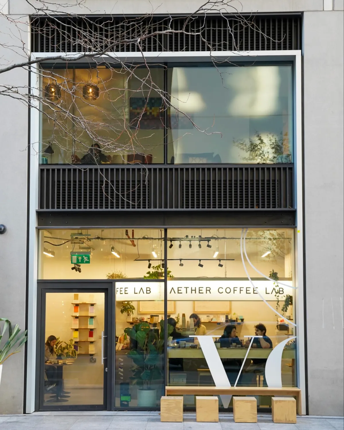 The glass front of Aether Coffee Lab in Wood Wharf, with the café name across the window, people at tables inside and wooden stools on the pavement
