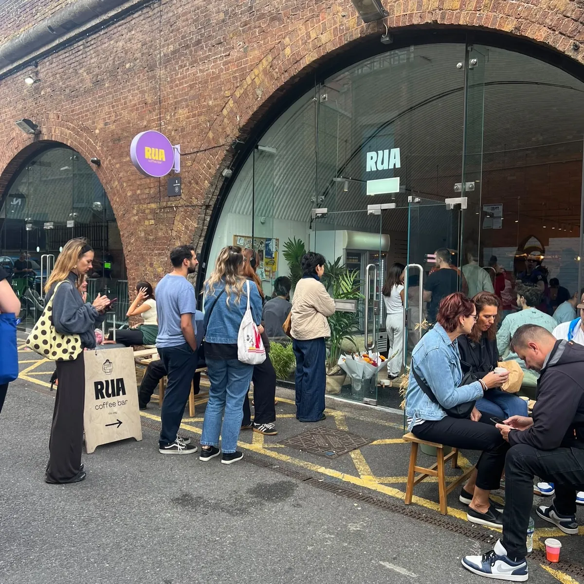 A crowd queuing and sitting on stools outside Rua's glass-fronted railway arch in Holloway, beside a Rua sandwich board