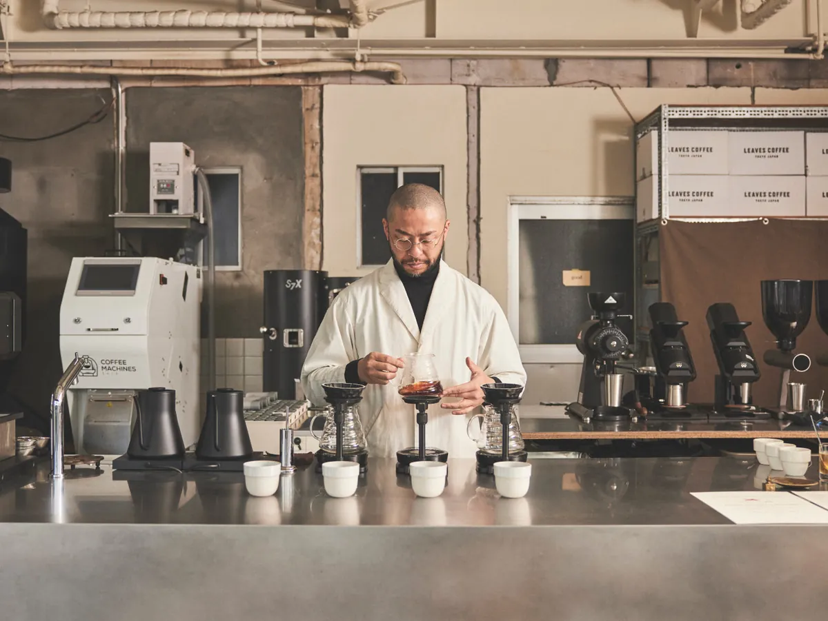 A barista in a white coat brewing pour-overs at the steel counter of LEAVES COFFEE ROASTERS, with a Stronghold roaster and grinders behind