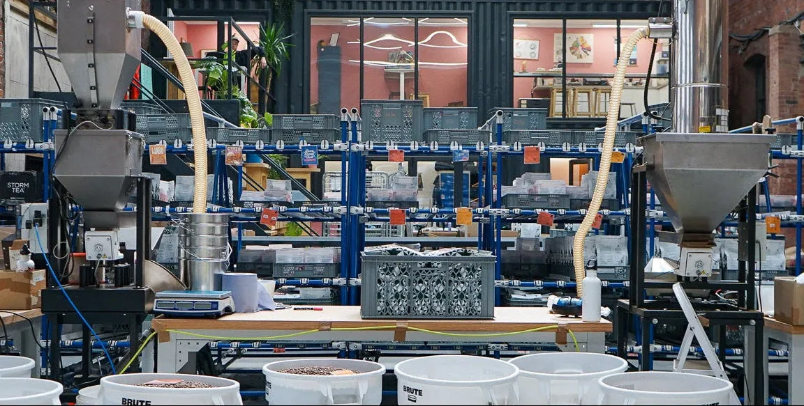 Roasting and packing equipment at the North Star roastery in Leeds, with hoppers, grey crates on blue shelving and white tubs of roasted coffee on the floor