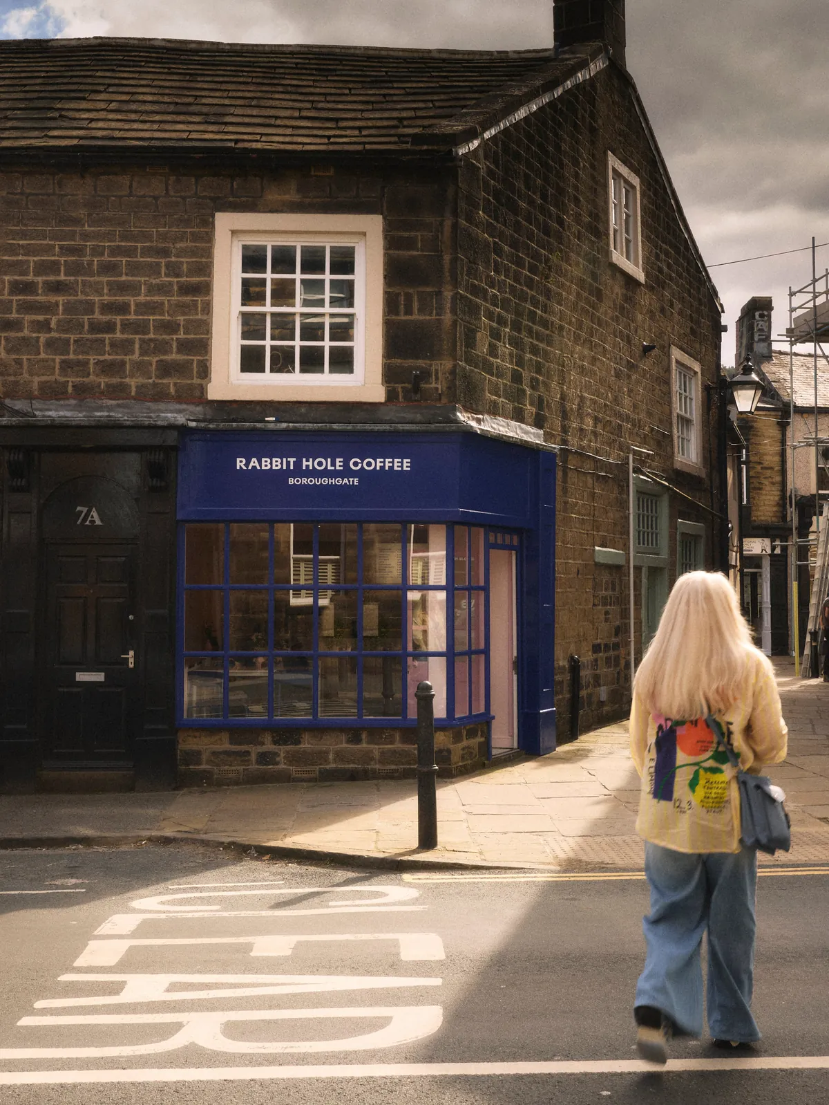 The stone corner building of Rabbit Hole Coffee in Otley, with a blue shopfront and a woman walking past on the street