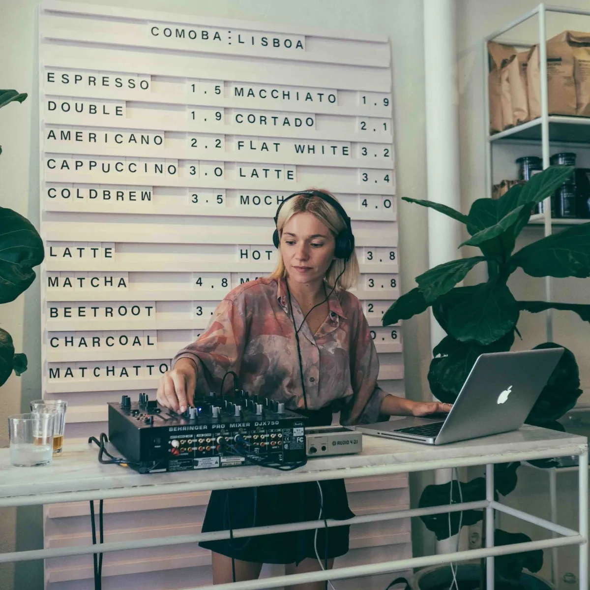 A DJ at a white marble table in front of Comobå's letter-board drinks menu, between large potted plants and white shelving