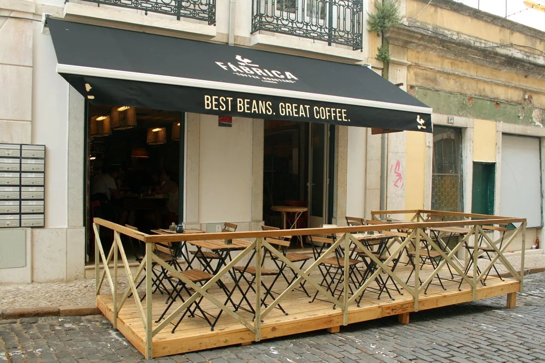 The black Fábrica awning reading Best Beans. Great Coffee. over the open doors of the café, with a wooden terrace of folding chairs on the cobbled street