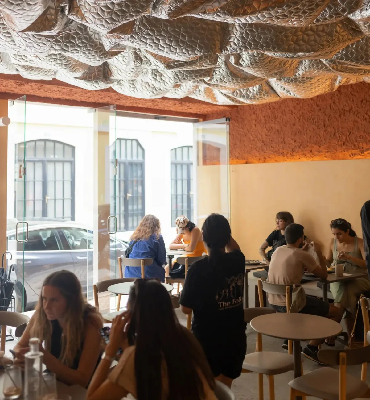 Guests at round tables in The Folks' first café in Baixa-Chiado, under a quilted silver ceiling and an orange textured wall, with glass doors open to the street