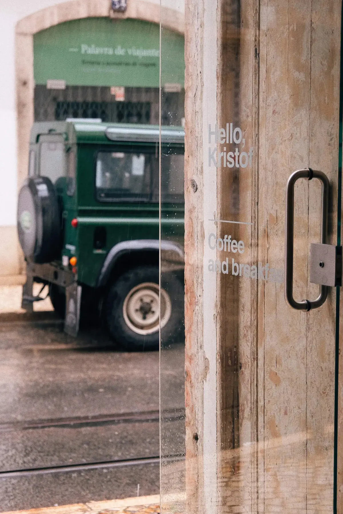 The glass door of Hello Kristof lettered Coffee and breakfast, with a green Land Rover parked on the tram-lined street outside