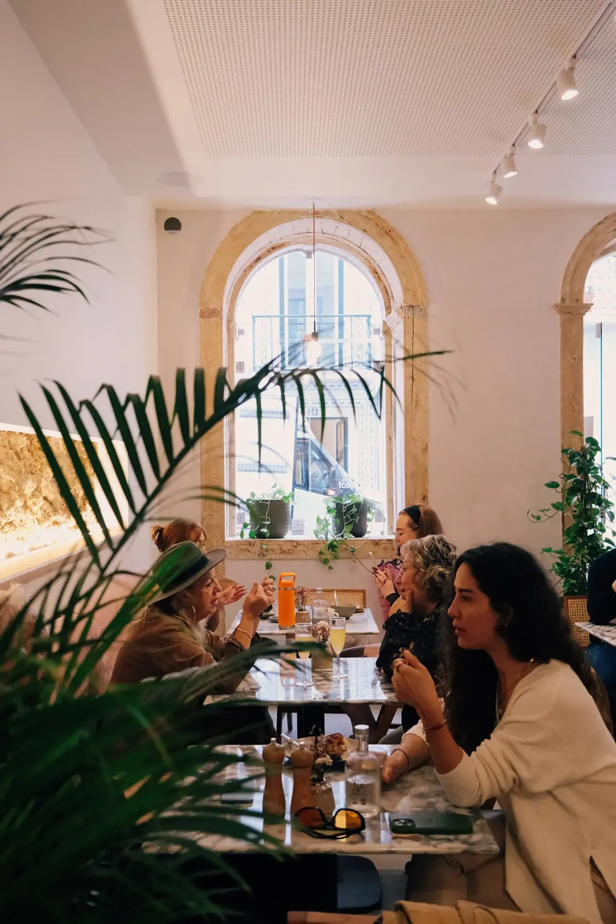 Guests at marble tables beneath a tall arched window in Hello Kristof's São Bento café, with palm leaves in the foreground