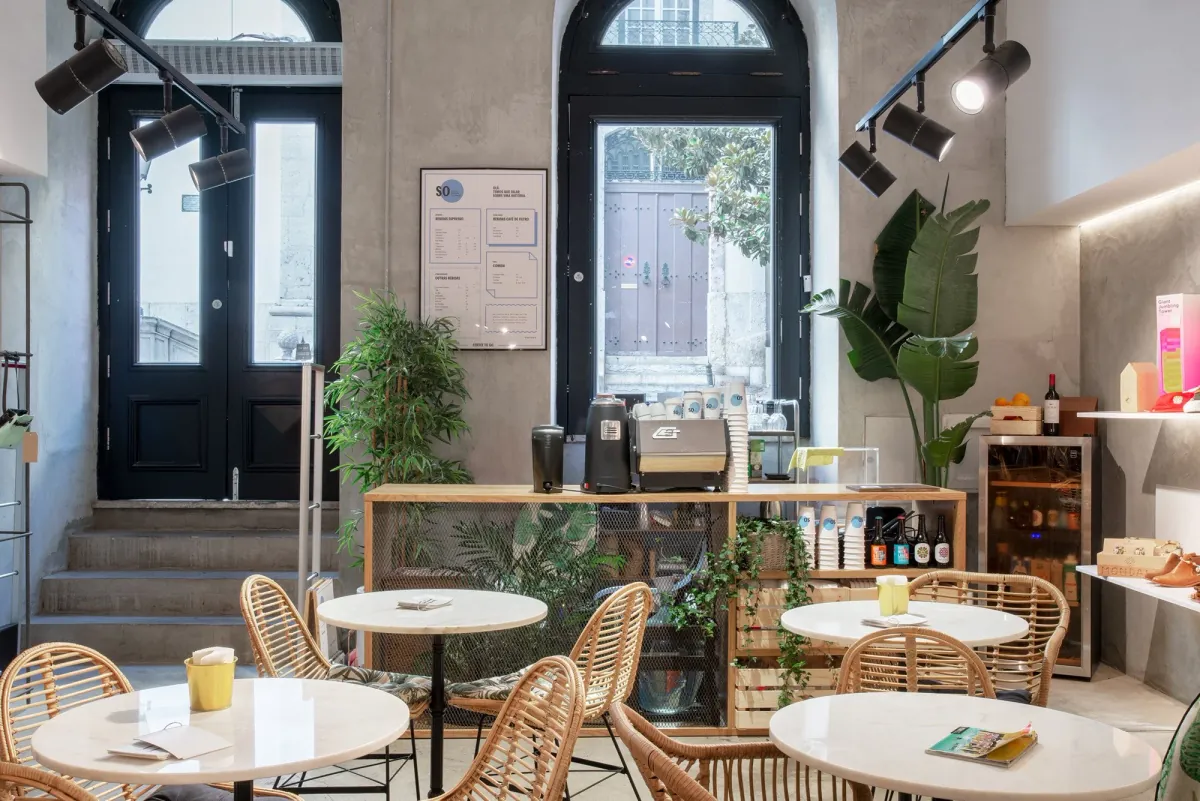 The SO Coffee Roasters bar in Lisbon, a wooden counter with an espresso machine and grinders below two tall arched doors, with rattan chairs and marble tables in front