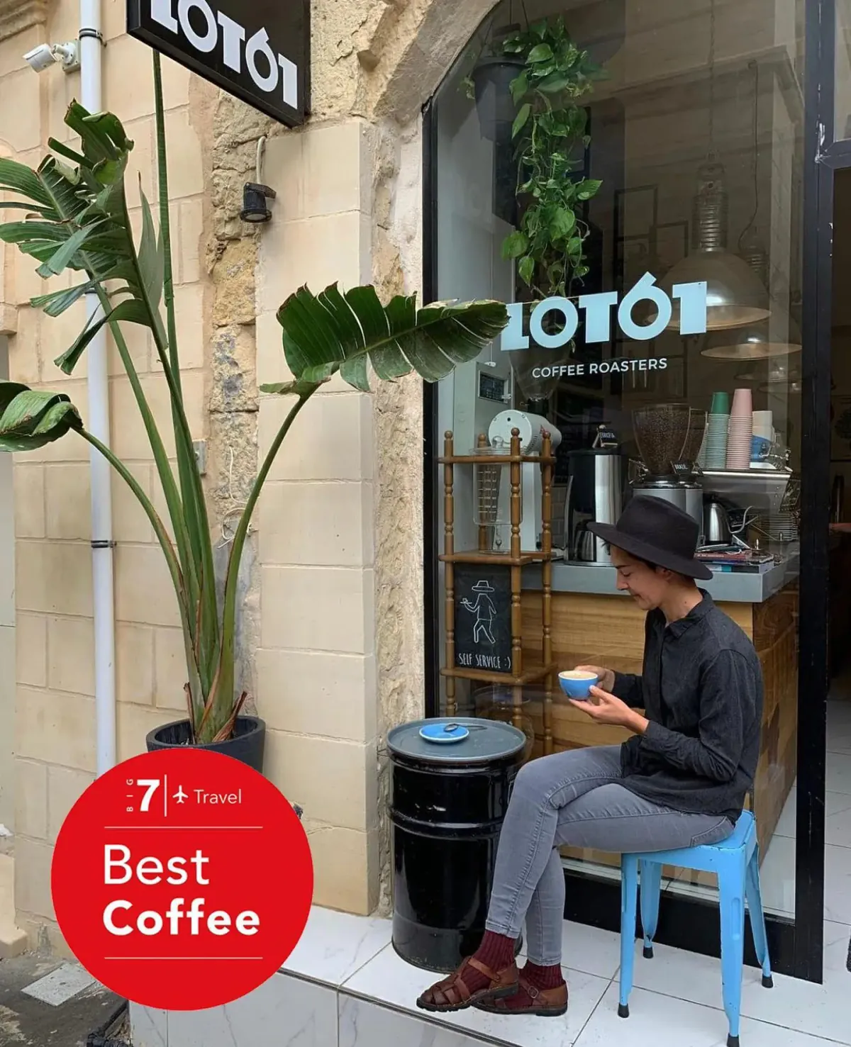 A customer in a black hat sits on a blue stool with a cup outside the glass front of the LOT61 coffee bar in Valletta, beside a potted plant, with a Big 7 Travel badge in the corner