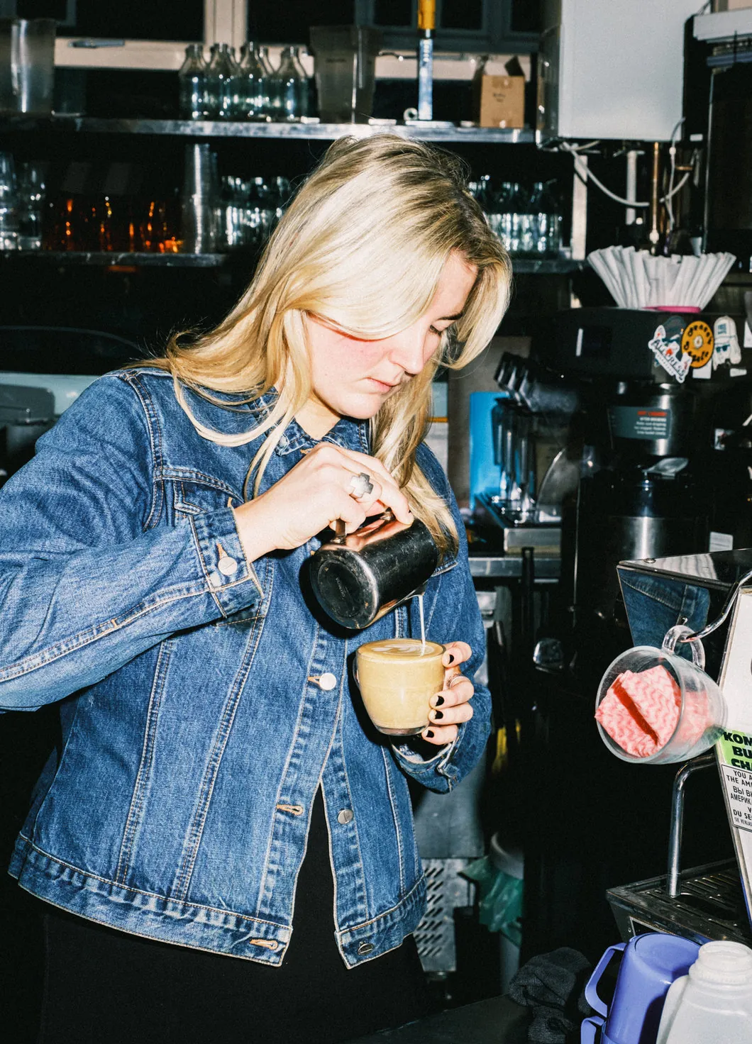 A barista in a denim jacket pouring steamed milk into a cup by the espresso machine at Milk