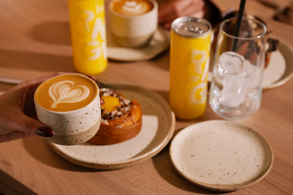 A hand holds a latte with a heart in the foam above a pastry on a speckled plate, with cans and an iced drink on a wooden table