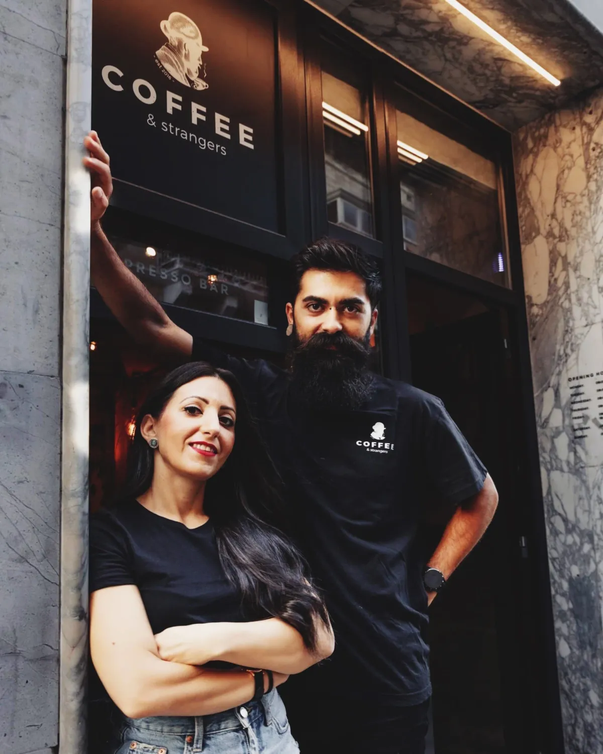 Two baristas in black COFFEE & strangers T-shirts at the door of the espresso bar in Sliema, under the sign with the bearded-stranger logo