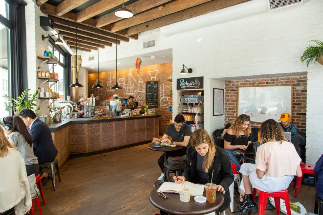 People reading and working at small tables in a sunlit Partners Coffee café with wooden beams, white-painted brick and a tiled counter