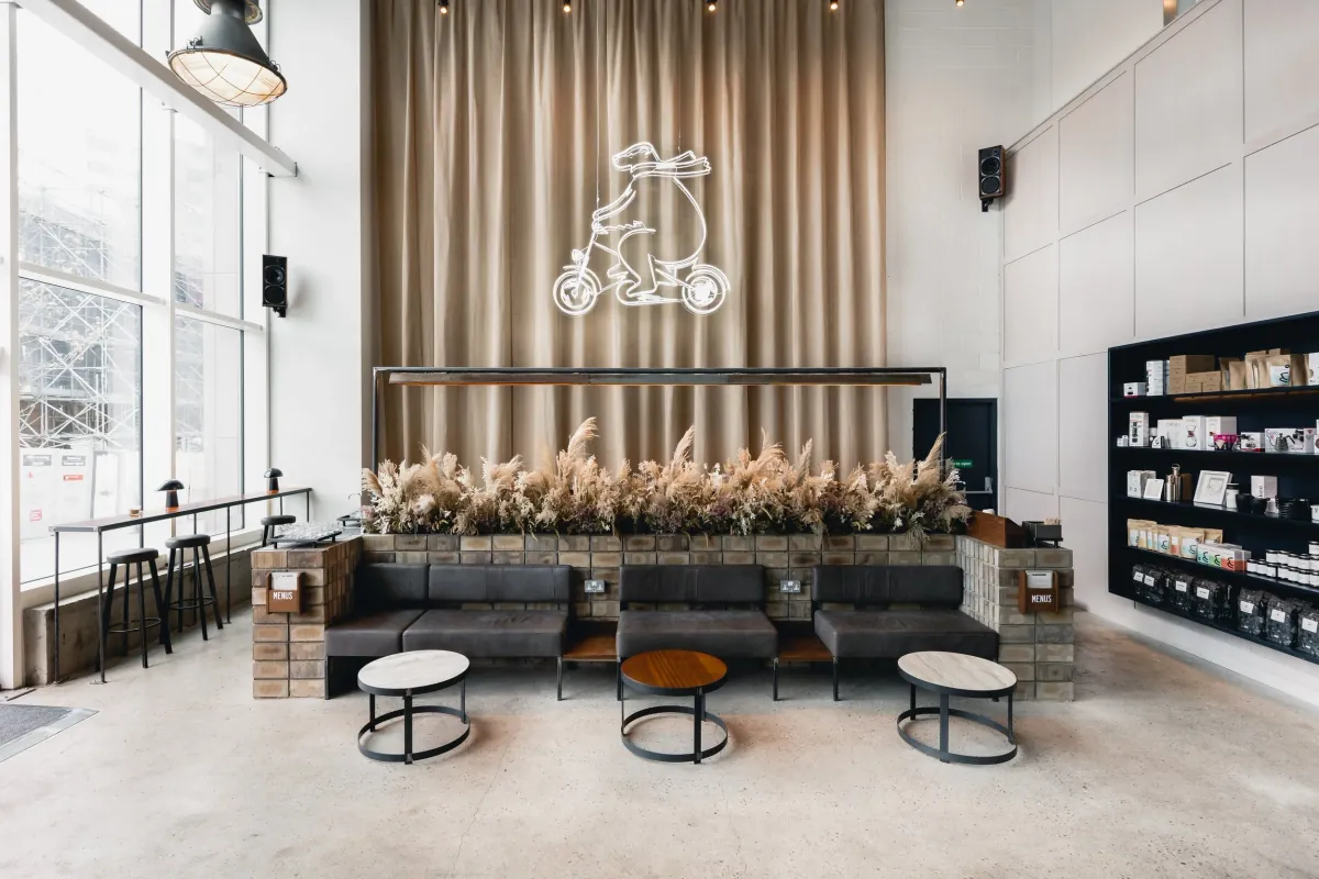 The tall glass-walled room of Marmadukes' coffee flagship in Sheffield city centre, with a long sofa, round tables, dried pampas grass and a neon bear on the curtained wall