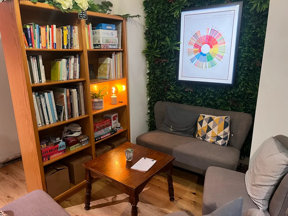 A corner of the downstairs room at Stage in Leeds, with grey sofas, a low wooden table, a bookcase of books and board games and a coffee flavour wheel on a green wall