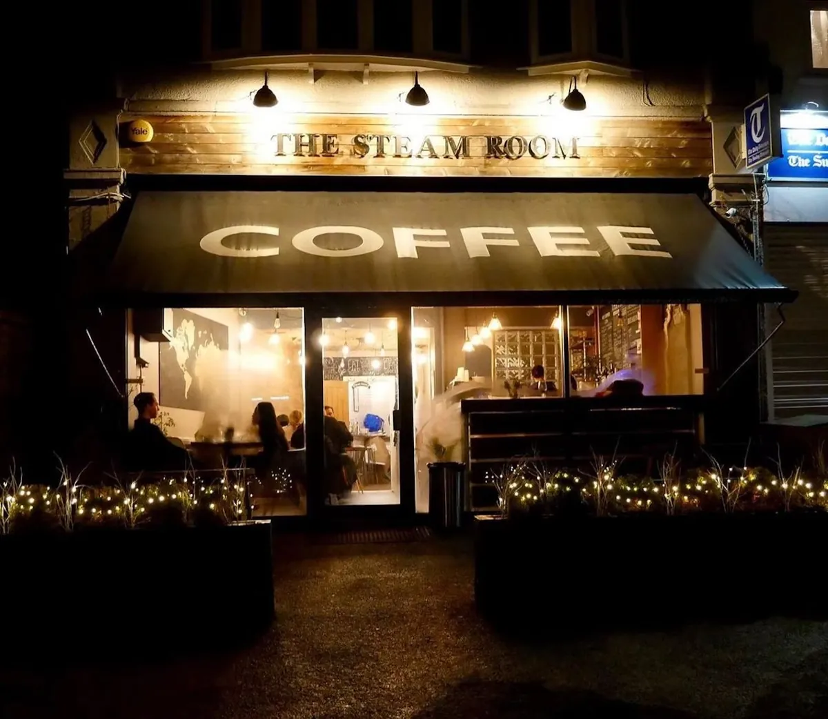 The Steam Room in Harborne at night, with the name lit above a dark awning reading Coffee, warm light through the glass front and fairy lights along the planters