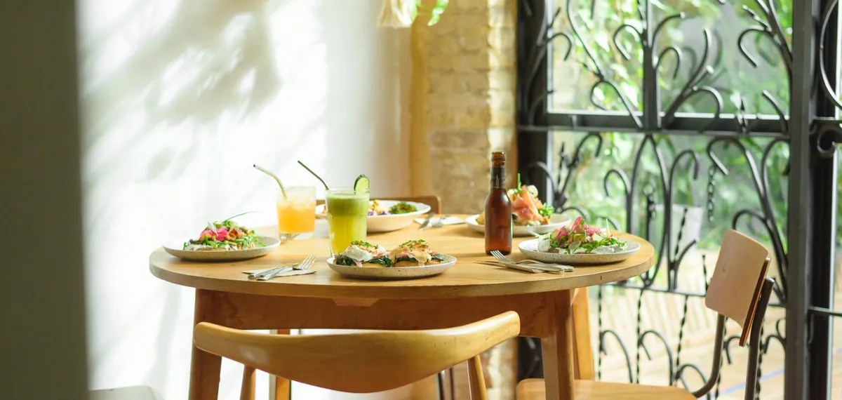 A round wooden table set with brunch plates, juices and a bottle beside a tall wrought-iron window at Bluebell Russafa