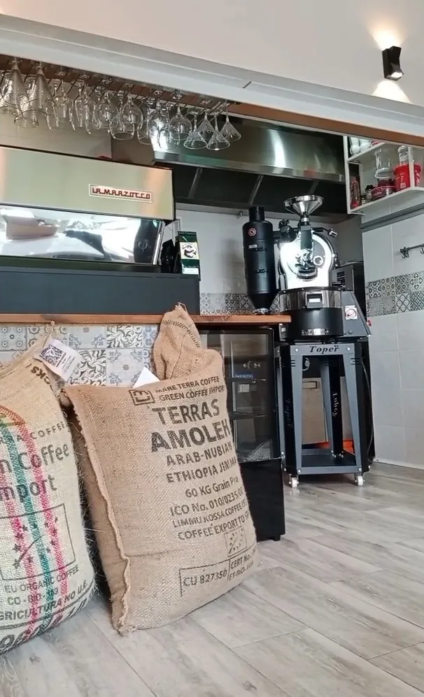 Jute sacks of green coffee in front of the black Toper roaster and the espresso machine inside Café Colomer