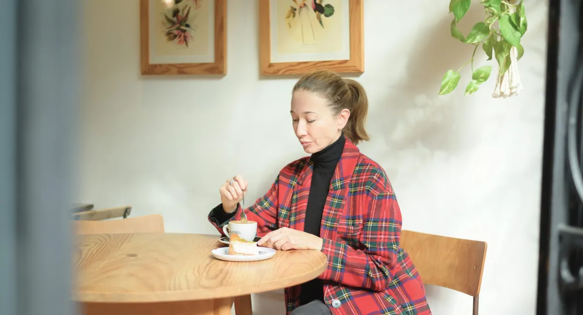 A woman in a red tartan jacket stirs a cup of coffee beside a slice of cake at a wooden table in Bluebell Russafa, under framed botanical prints