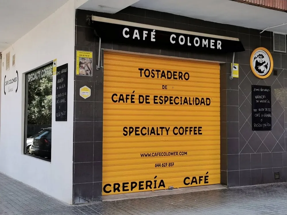 The front of Café Colomer in Valencia, with a yellow shutter painted Tostadero de café de especialidad and Crepería Café under a black awning