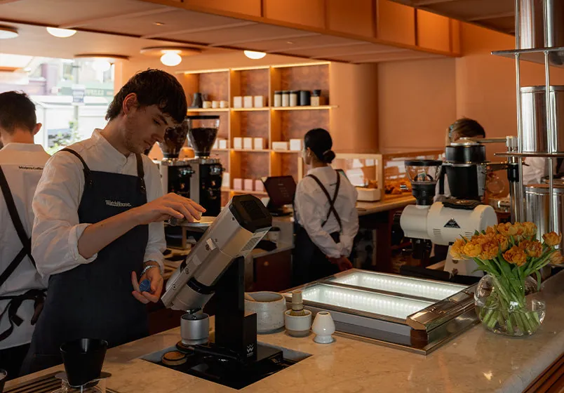 Baristas brewing behind a WatchHouse bar, with grinders, a filter station and retail shelves