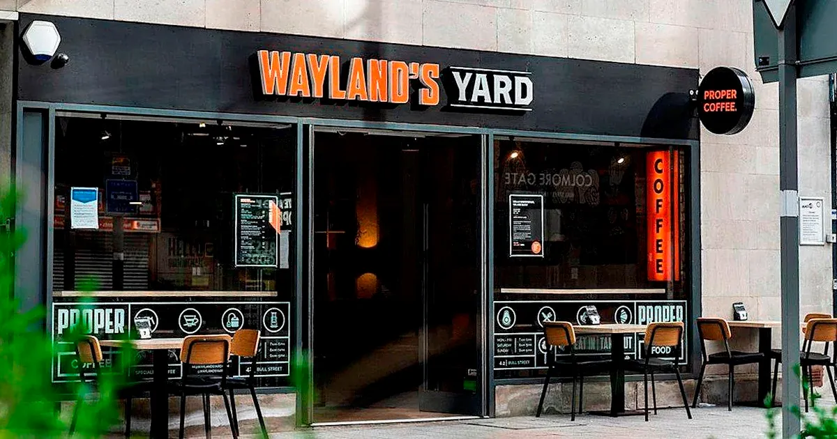 The glass shopfront of Wayland's Yard in Birmingham, with its orange and white name sign, a Proper Coffee roundel and wooden chairs outside